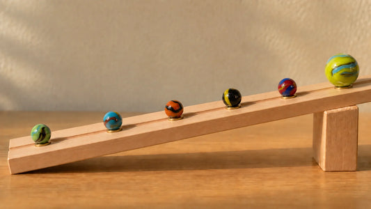 Planche de bois inclinée posée sur un bloc, six billes de verre MesBilles retenues chacune par un petit repère de laiton, dont les écarts s'élargissent à mesure que l'on descend la pente